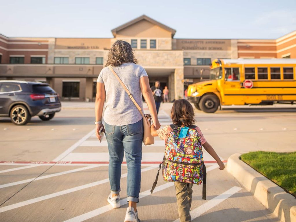 A parent holding a kids hand and walking through the parking towards the school entrance.